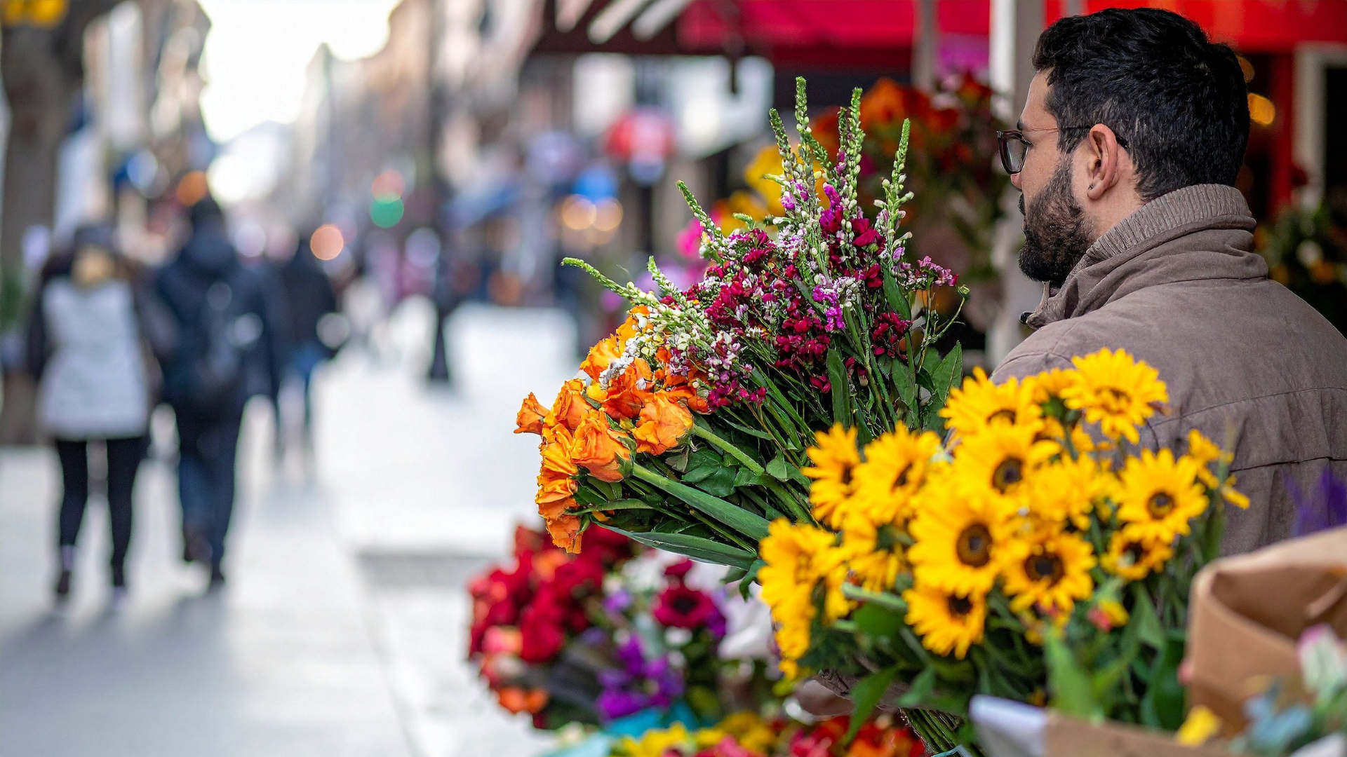 Uomo che vende fiori su una strada affollata, circondato da piante colorate e passanti interessati.